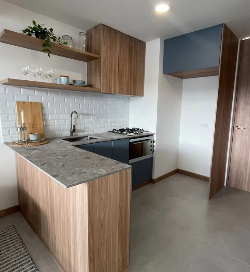 Cocina del apartamento modelo Quibay: muebles en madera y azul, mesón en piedra gris y repisas flotantes